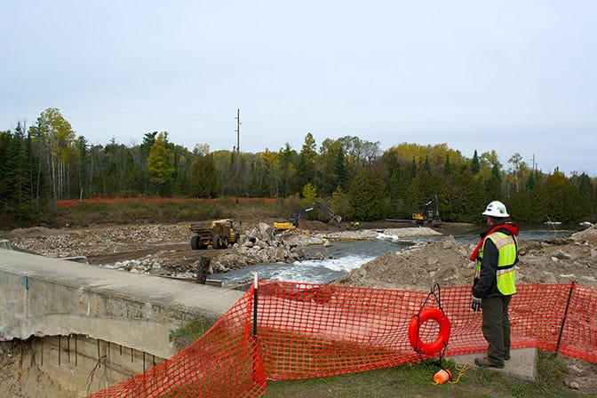 A person in reflective vest and hard hat stands atop a dam