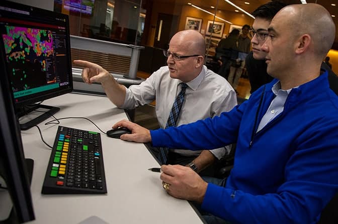 Three people gather around a computer, with one pointing at the screen.