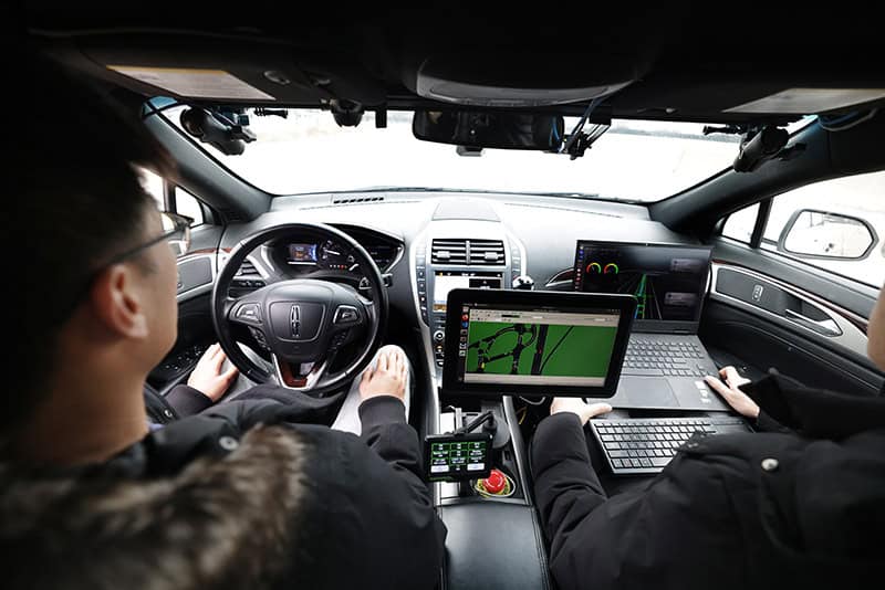 Two researchers are seen from behind, sitting in a test vehicle. The passenger works on a laptop computer while a monitor screen sits on the console between them.